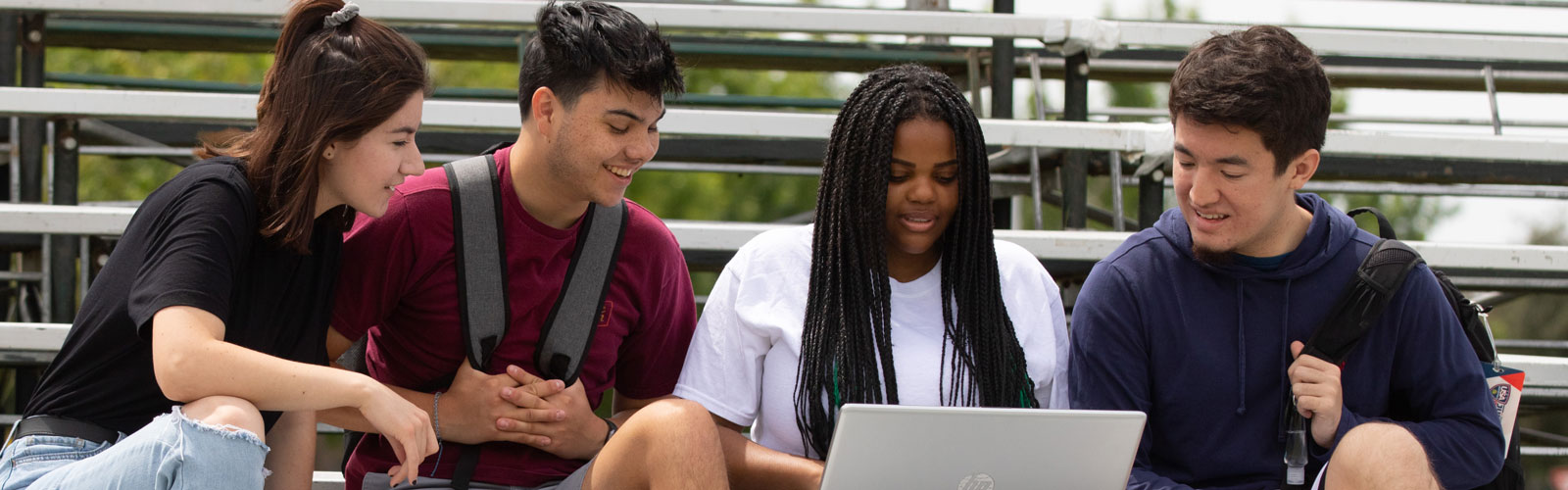 4 students sitting on bleachers looking at laptop and smiling