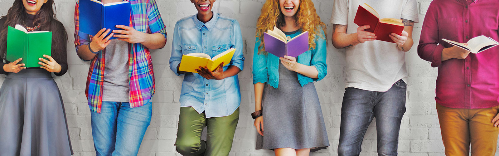 a row of young people of different ethnicities holding books and smiling while standing against a white brick wall.