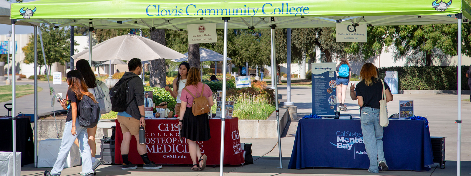 Transfer day festival, people talk to representatives from Cal State Monterey and CHSU while under the shade of a canopy that says "Clovis Community College"
