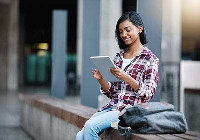 a student with dark hair in a purple plaid shirt is sitting on a wall looking at a tablet.