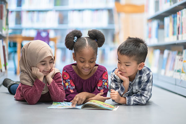 a little girl wearing a hajib, a little black girl, and a Chinese boy laying on the floor of a children's library reading a book