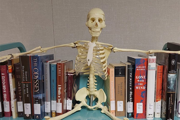 A skeleton model sits on a shelf of challenged books